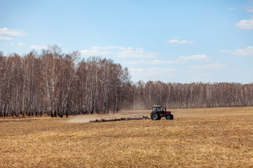 Obraz premium Red tractor with a trailed plow for mowing and weeding fields for the agro-industry of yellow color under the blue sky, a clear spring day. Preparation for planting crops rural technology. Farming.