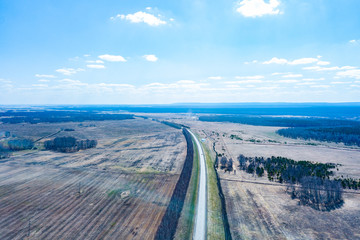 Aerial view of the asphalt road in a field with yellow grass during planting seeds and crops far from big cities on a clear spring day under a blue sky with trees to protect from the wind.