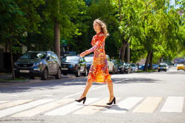 Young beautiful blonde woman in a red flower dress crosses the road at a crosswalk