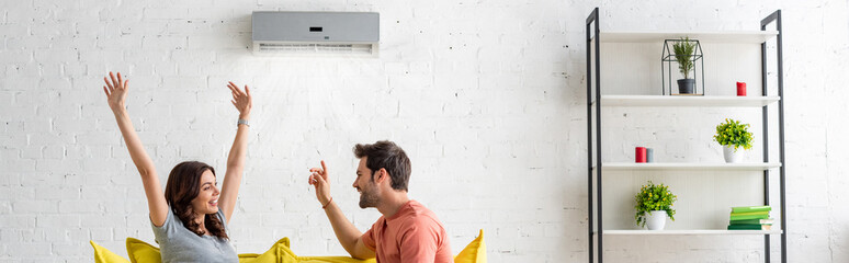 Fototapeta panoramic shot of excited man and woman sitting under air conditioner at home obraz