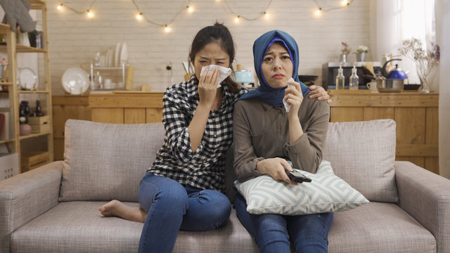 Multiracial young people watching sad movie in TV relax on sofa with home kitchen in background in bright cozy apartment. two asian chinese and muslim women friends upset crying using napkin tissue