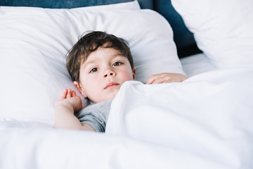 cute kid lying on white pillow in bed and looking at camera