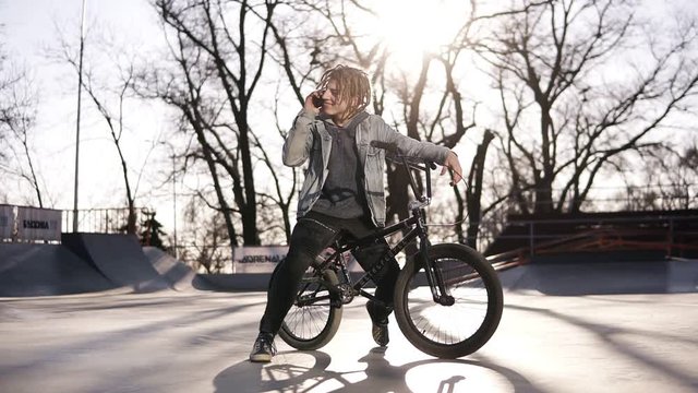 Portrait of happy handsome teenage boy with dreadlocks, using mobile phone while riding a BMX bike, smiling, messaging friends via social networks. People, lifestyle and communication concept. Low