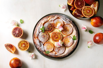 Homemade Cheesecake with sicilian blood oranges, decorated by edible flowers, mint leaves and sugar powder served in plate with cutted oranges above over white marble background. Flat lay, space