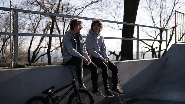 Two teenage men are sitting on high parapet together. Bmx rider and skateboarder - a boy with the dreadlocks is openly smoking. Bad habit. Street life
