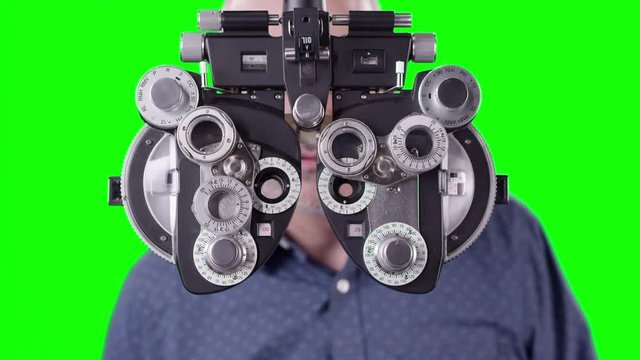Man walks behind and looks through Phoropter for eye exam against green screen for editing.