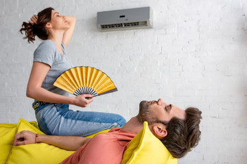 young woman with hand fan sitting on yellow sofa under air conditioner near lying man - obrazy, fototapety, plakaty