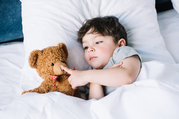 adorable kid lying on white pillow and touching teddy bear at home