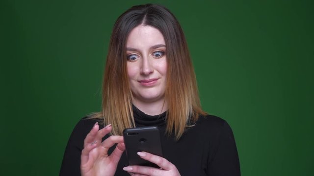 Young businesswoman with chestnut hair watches into cellphone in perplexity and confusion on green background.