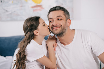 happy daughter kissing cheek of cheerful father in bedroom