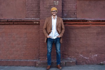 Obraz premium Portrait of stylish handsome young man in glasses with bristle standing outdoors. Man wearing jacket and shirt, leaning against wall.