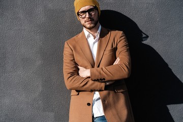 Portrait of stylish handsome young man in glasses with bristle standing outdoors. Man wearing jacket and shirt, leaning against wall.