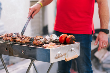 Handsome athletic man cooking barbecue outdoors. Brutal man with jeans and red shirt standing and prepared meat and vegetables on a courtyard.