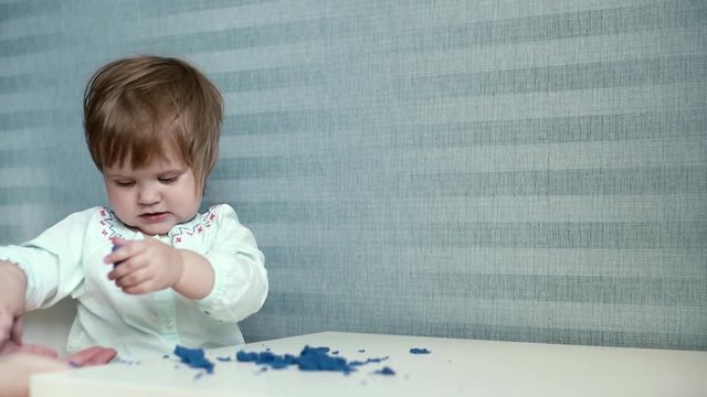 Little cute baby girl sits at a childs table and dumps kinetic sand from the table to the floor in the background of wallpaper in slow motion. Loving mom picks up kinite sand that throws her daughter