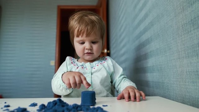 Little cute baby girl sits at a childs table and plays with kinetic sand on the background of beautiful blue wallpaper in slow motion. Kid thinks for a long time before breaking a kinetic sand figure