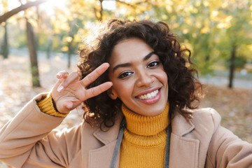 Obraz premium Young african happy woman walking outdoors in a spring park take a selfie by camera make peace gesture.
