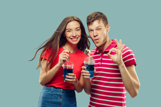 Beautiful young couple's half-length portrait isolated on blue studio background. Woman and man standing with drinks smiling and signing OK. Facial expression, summer, weekend concept. Trendy colors.