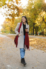 Portrait of stylish brunette woman wearing coat drinking takeaway coffee and smiling in autumn park