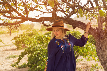 Beautiful blond hair woman wearing blue skirt and brown hat near tree in vineyard - plantation of grapevines. Spring dawn solar bright warm effect. Slow living lifestyle. Local agriculture business.