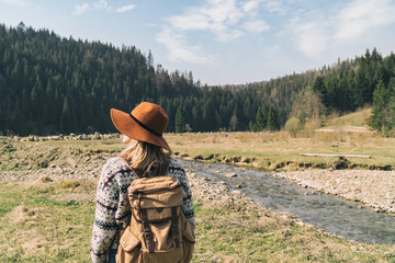 Stylish hipster wearing brown hat and wool sweater with vintage textile backpack near mountains river and forest. Blond hair woman exploring nature. Travel and wanderlust concept. Amazing atmospheric.