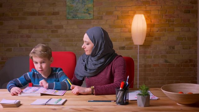 Muslim mother in hijab learning letters with her small son at the table at home.