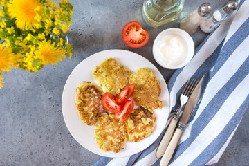 fritters of zucchini with slices of tomato on the plate, gravy boat with cream sauce, a fork,  knife,  towel,  bouquet of dandelions, salt, pepper, jar of oil, top view,