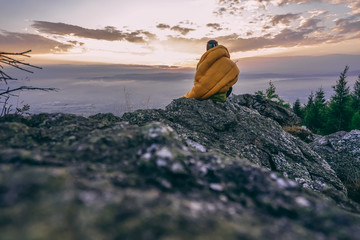 A man siting on a rock with his sleeping bag and watching sunrise or sunset. Beautiful outdoor shot, mountain landscape and dramatic sky, a man in a sleeping bag camping in the outdoors.
