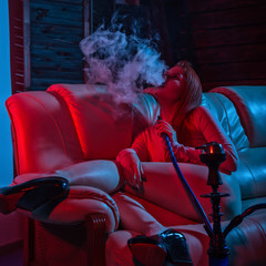 Sexy redhead woman vaping electronic hookah in red blue tones. girl blows white doom