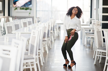 Obraz premium Сheerful business african american lady with afro hair, wear white blouse and green pants posed in cafe.