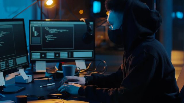 cybercrime, hacking and technology concept - asian male hacker in mask writing code or using computer virus program for cyber attack in dark room at night
