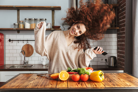 Image of happy beautiful woman listening to music on mobile phone while cooking at home