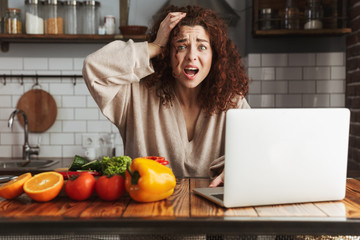 Image of nervous caucasian woman using laptop while cooking in kitchen interior at home