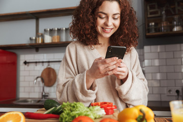 Photo of happy caucasian woman holding smartphone while cooking salad with fresh vegetables at home