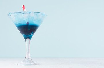 Fresh blue alcohol cocktail with ice cubes, red straw in misted wineglass on green color background, white wood board.