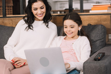 cheerful mother looking at laptop near daughter listening music in earphones