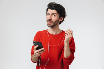 Handsome young serious man posing isolated over white wall background listening music with earphones using mobile phone.