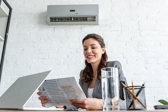 smiling businesswoman reading newspaper while sitting at workplace under air conditioner
