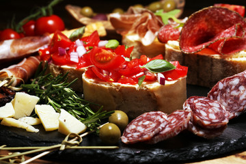 Traditional italian antipasto bruschetta appetizer with cherry tomatoes, cream cheese, basil leaves and balsamic vinegar on cutting board with prosciutto, salami, cheese