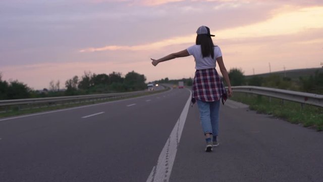 Slow-motion rear view hipster woman tourist walking forward highway countryside road, evening dawn, car broke down, stretch hand thumb up hitchhiking try find car give ride, lift to destination.