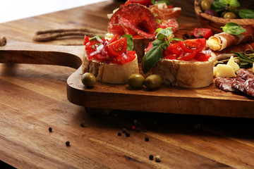 Traditional italian antipasto bruschetta appetizer with cherry tomatoes, cream cheese, basil leaves and balsamic vinegar on cutting board with prosciutto, salami, cheese