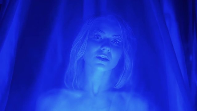 Portrait of young sexy woman with snow makeup on her eyelashes, posing under tulle erotical, blue light, winter night scene, close-up