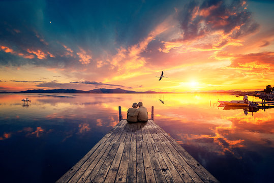 Senior couple seated on a wooden jetty, looking a colorful sunset on the sea with a flying flamingo reflected on the calm water.
