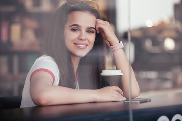 View through the glass portrait of a beautiful young cheerful girl sitting at a table in a cafe with a cup of hot coffee and a smartphone. Concept of wireless internet and leisure in the city