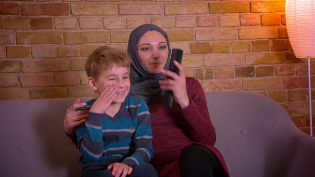 Portrait of small boy and his muslim mother in hijab hugging and watching TV together sitting on sofa at home.