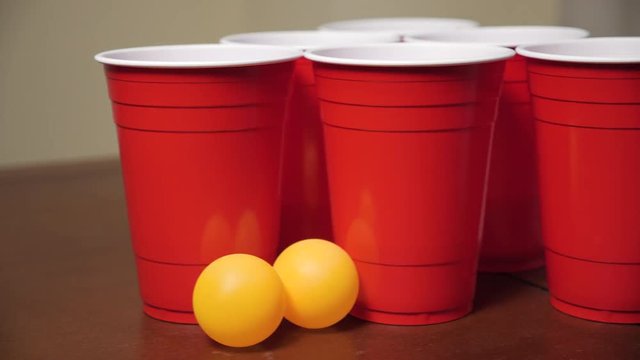 A group of red plastic cups and orange ping pong balls sit on a table, ready for a game of beer pong