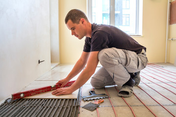 Young worker tiler installing ceramic tiles using lever on cement floor with heating red electrical...