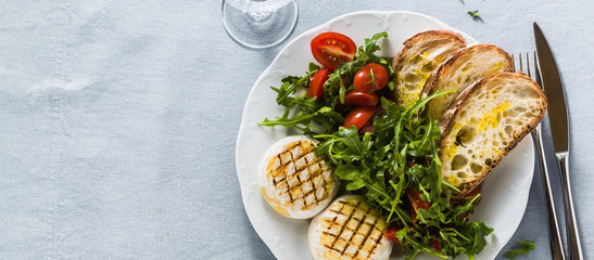 banner of grilled Italian Tomino cheese served on a table with arugula salad and fresh homemade ciabatta bread and tomato on a blue linen festive tablecloth. summer menu