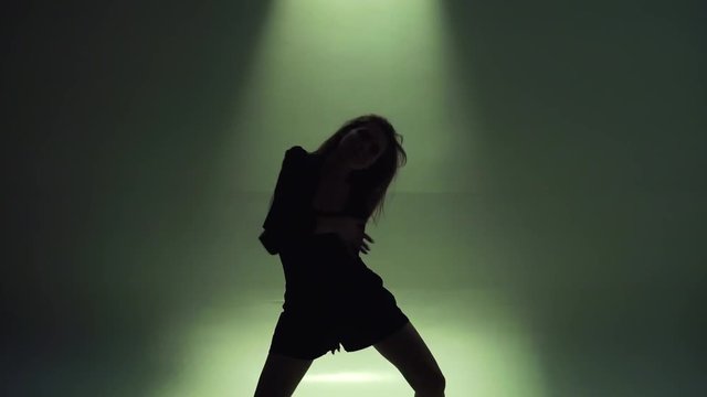 Silhouette of young woman with long hair in black clothes dancing erotic dance in green studio light, moving fast and emotionally