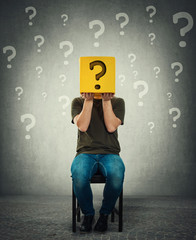 Incognito young man seated on a chair holding a yellow box with question mark instead of head.
