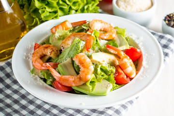 Fresh shrimp salad made of tomato, ruccola, avocado, prawns, chicken breast, arugula, crackers and spices. Caesar salad in a white, transparent bowl on wooden background